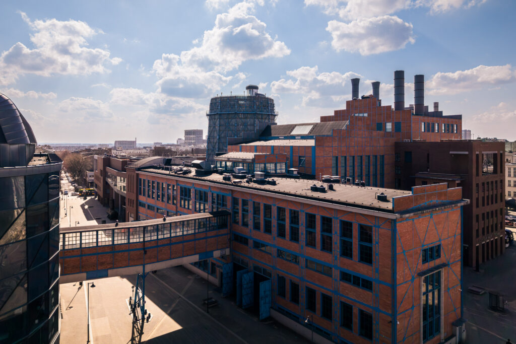 Zdjęcie przedstawia panoramę miejskiego kompleksu architektonicznego, charakteryzującego się ceglanymi fasadami z wyraźnymi, niebieskimi ramami okiennymi i elementami konstrukcyjnymi. Centralnym punktem kompozycji jest przeszklony most łączący dwa - Architektura, Dron Photography 51.768328, 19.469030