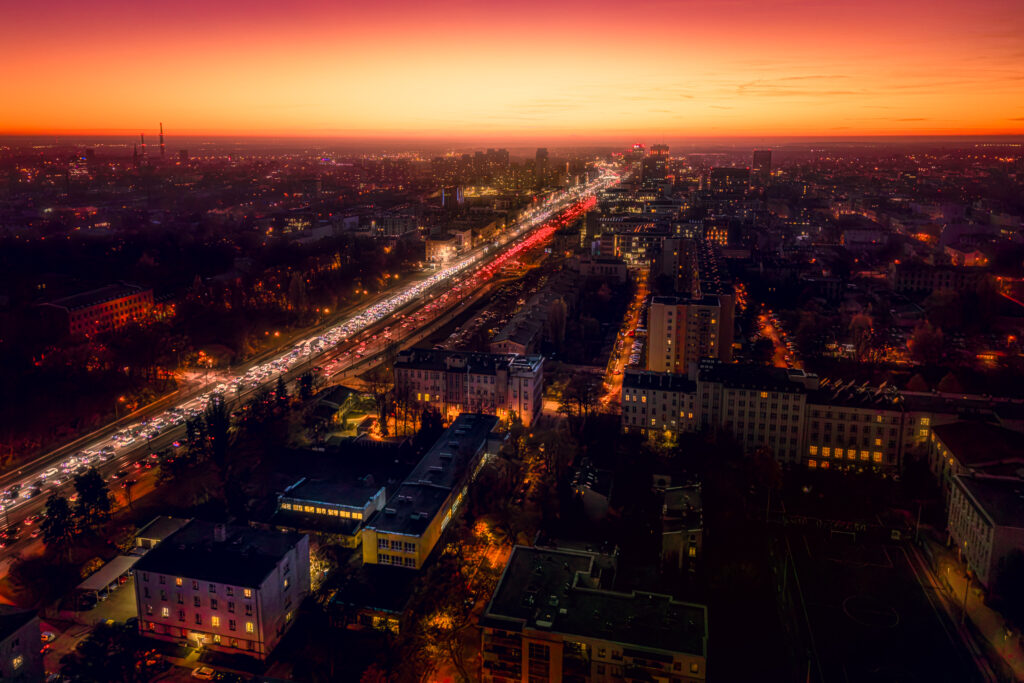 Fotografia lotnicza ukazuje rozległą panoramę miejską podczas zachodu słońca, charakteryzującą się dynamicznym ruchem ulicznym. Długa ekspozycja uchwyciła smugi świateł pojazdów, tworząc świetliste linie wzdłuż głównej arterii komunika - Architektura, Dron Photography 51.762560, 19.480083