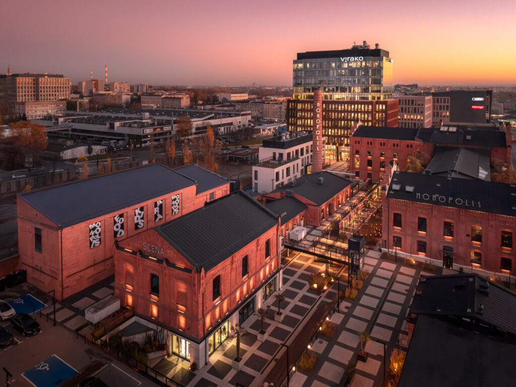 Panorama miejska przedstawiająca kompleks Monopolis w Łodzi, charakteryzujący się połączeniem historycznej architektury przemysłowej z nowoczesnymi elementami. Zdjęcie wykonane o zmierzchu ukazuje ciepłe barwy zachodzącego słońca, które podkr - Architektura Photography 51.764085, 19.484855